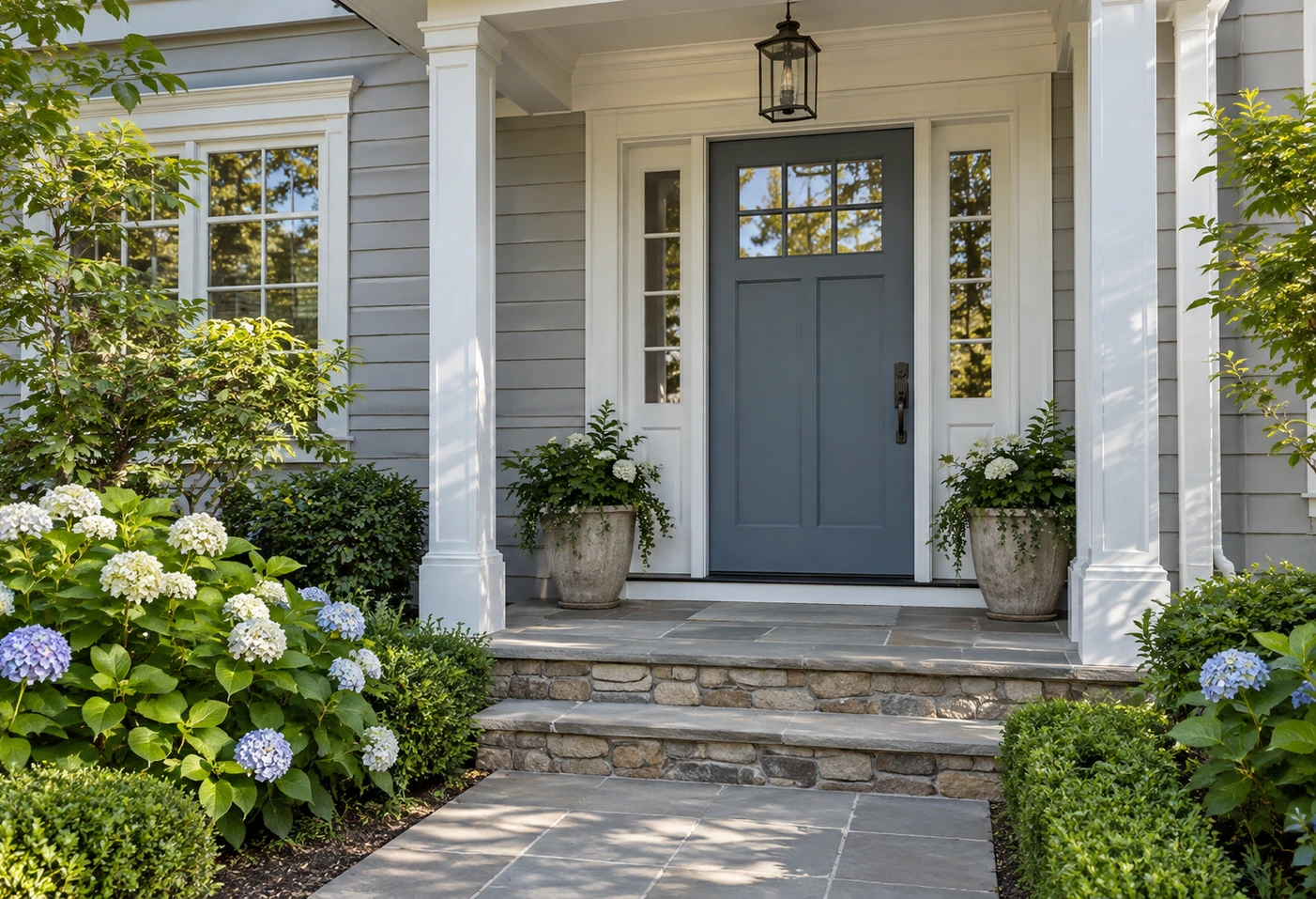 Blue-gray front door with white porch columns, gray siding, stone steps, and hydrangeas.