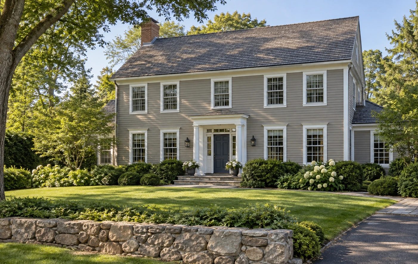 Gray colonial-style home with white trim, a dark front door, cedar shingle roof, landscaped lawn, and low stone wall.