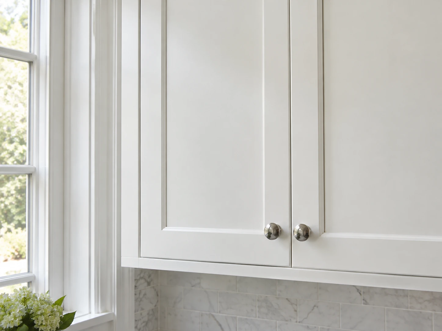 White painted kitchen cabinet doors with round metal knobs beside a bright window and marble tile backsplash.